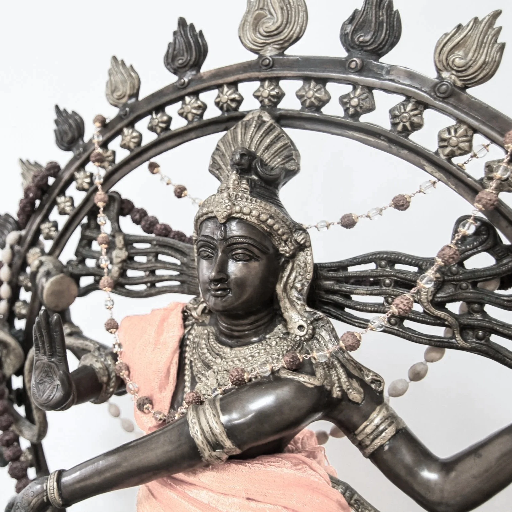 Silver-colored sculpture of a woman with traditional jewelry and a headdress, draped in pink cloth, with decorative elements and chains around her, set against a plain background.