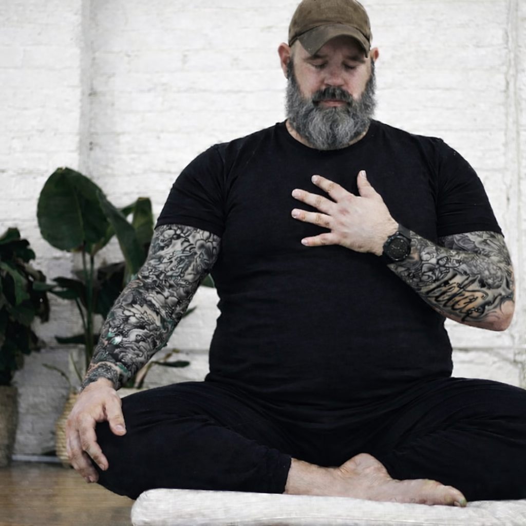 A man with a beard, tattoos, and wearing a black t-shirt and black pants is sitting cross-legged on the floor in a pose resembling meditation. He is wearing a brown cap, a watch, and is touching his chest with his right hand while resting his left hand on his knee.