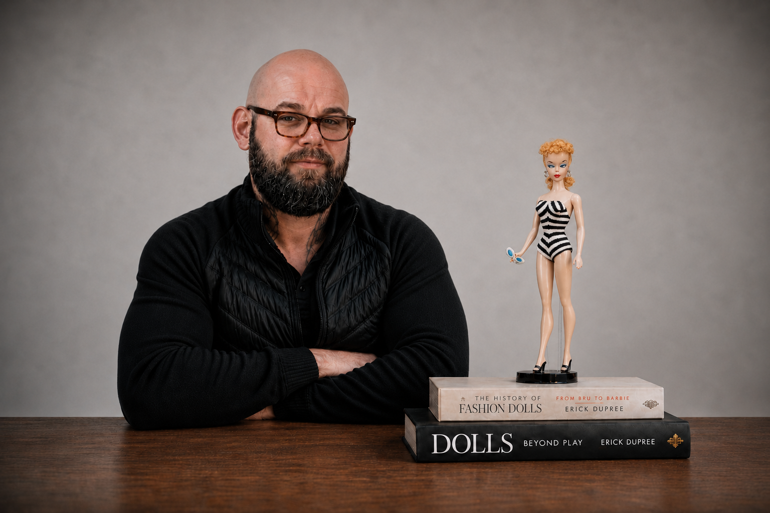 A man with glasses and a beard sitting at a wooden table with his arms crossed, next to a stack of two large books about dolls, with a fashion doll in a black and white striped swimsuit standing on top of the books. The background is plain gray.