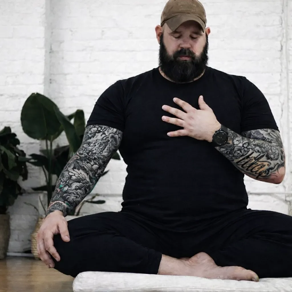 A man with a beard and tattoos wearing a camouflage cap and black clothing, sitting cross-legged indoors, with his right hand on his chest and eyes closed.