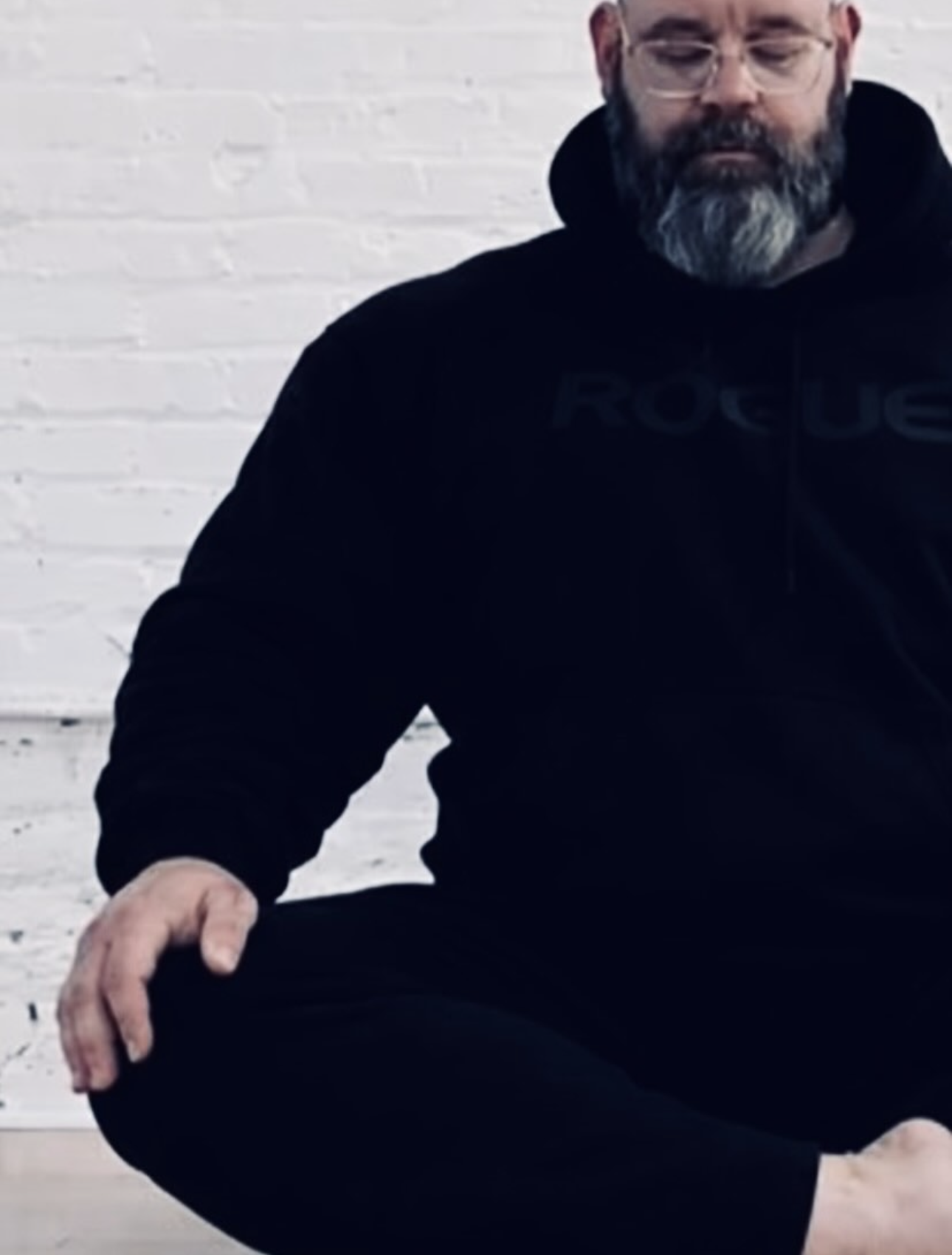 A man with glasses and a beard in a black hoodie sitting on the ground against a white brick wall.