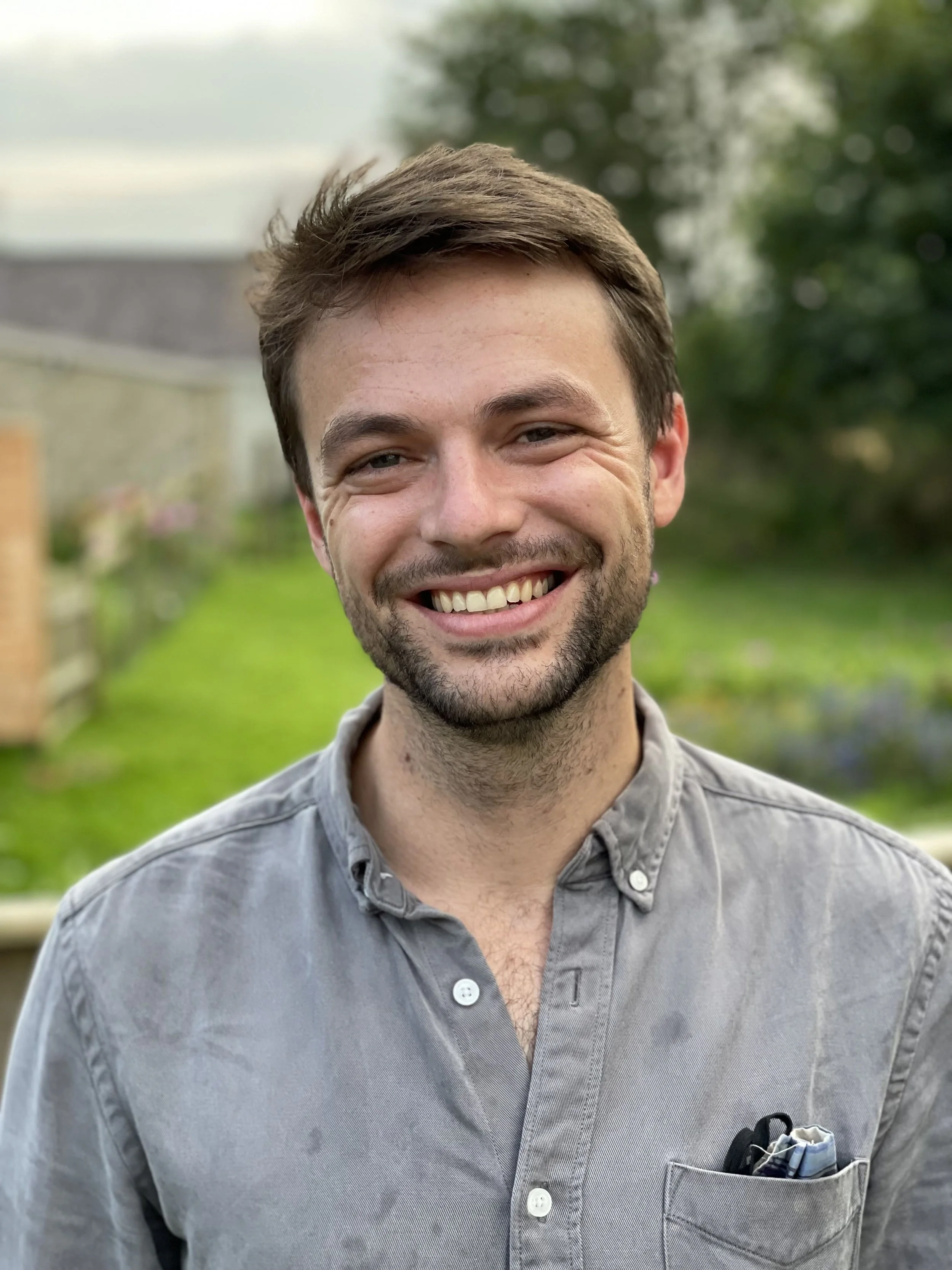 Dom Blake standing in a garden, smiling, with trees and grass in the background.