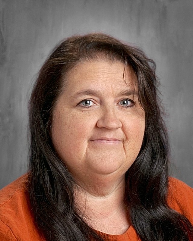 A woman with long dark hair, light skin, and blue eyes, wearing an orange top, posing in front of a gray background.