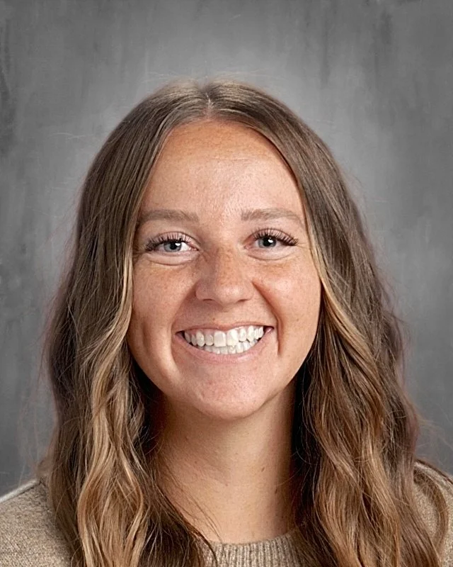 Close-up of a smiling young woman with long, wavy brown hair, wearing a beige top, and standing against a gray background.
