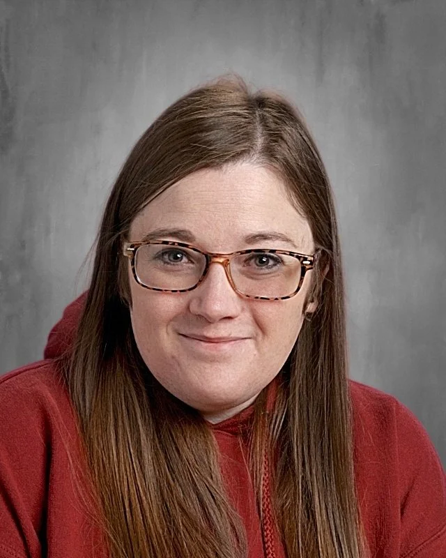 Portrait of a woman with brown hair, glasses, smiling, wearing a red hoodie, against a gray background.