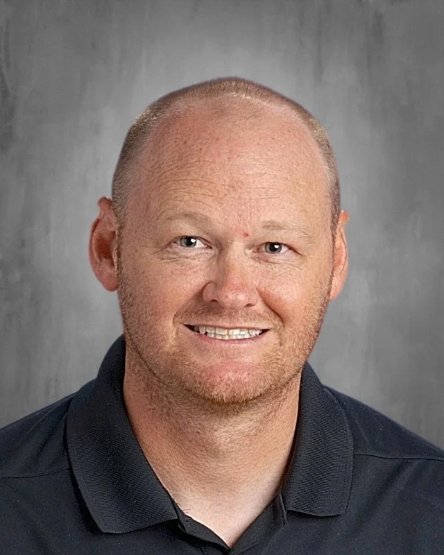 Headshot of a smiling man with short, light-colored hair, wearing a black collared shirt, against a gray background.