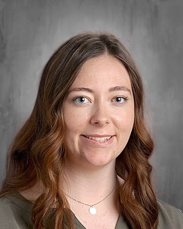 A portrait of a young woman with long, wavy brown hair and blue eyes, smiling softly, wearing a gray top and a delicate silver necklace, against a gray background.