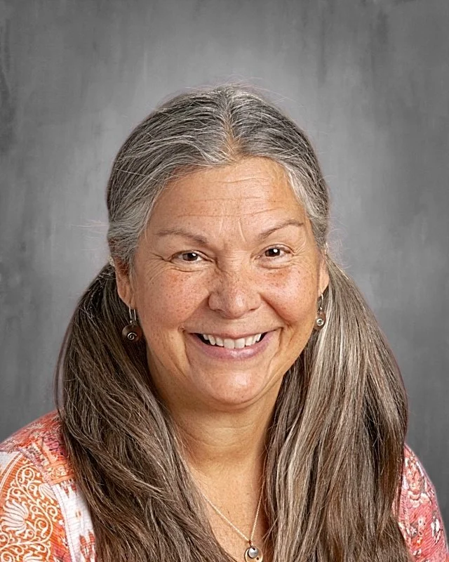 Smiling middle-aged woman with long gray hair and earrings, wearing a patterned top and necklace, against a gray background.