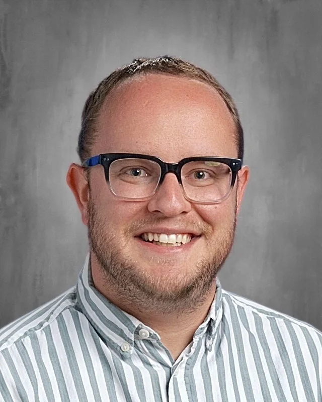 A smiling man with glasses, wearing a striped button-up shirt, standing against a gray background.