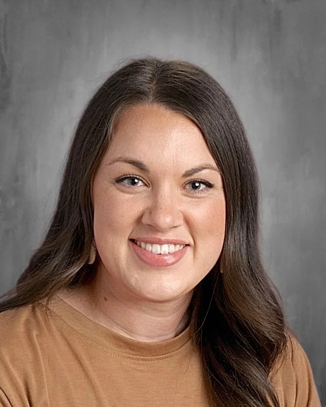 Close-up photo of a smiling woman with long brown hair and blue eyes, wearing a beige top, against a gray background.