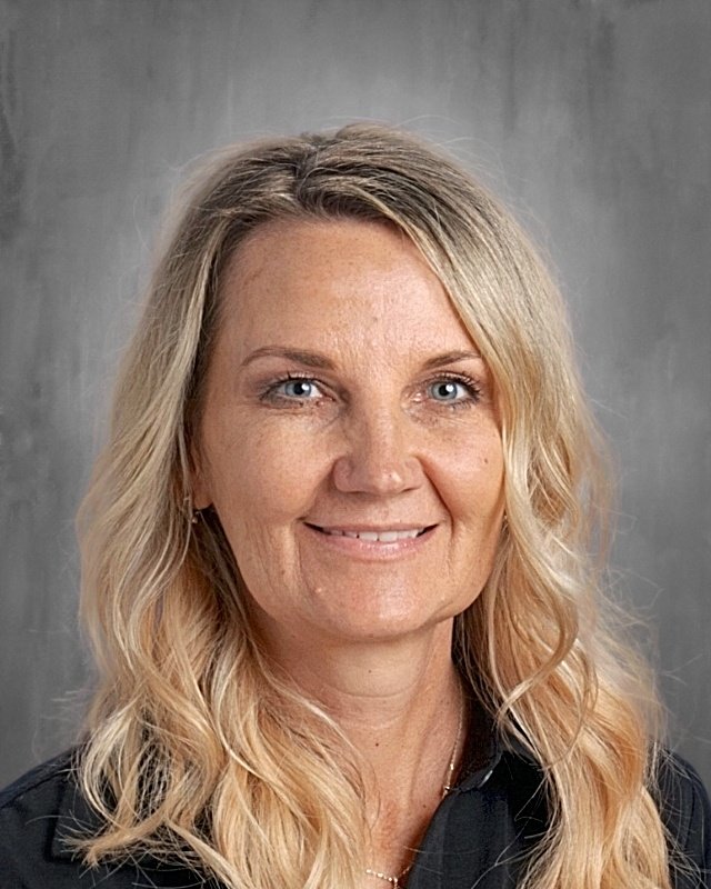 A professional portrait of a woman with shoulder-length wavy blonde hair, blue eyes, and fair skin, wearing a black top, smiling against a gray background.