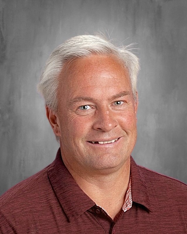 A middle-aged man with white hair and blue eyes, smiling, wearing a maroon collared shirt, with a plain gray background.