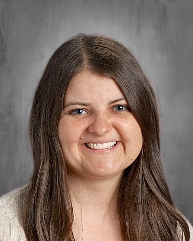 Close-up portrait of a smiling woman with long brown hair and blue eyes, wearing a neutral-colored top, against a gray background.