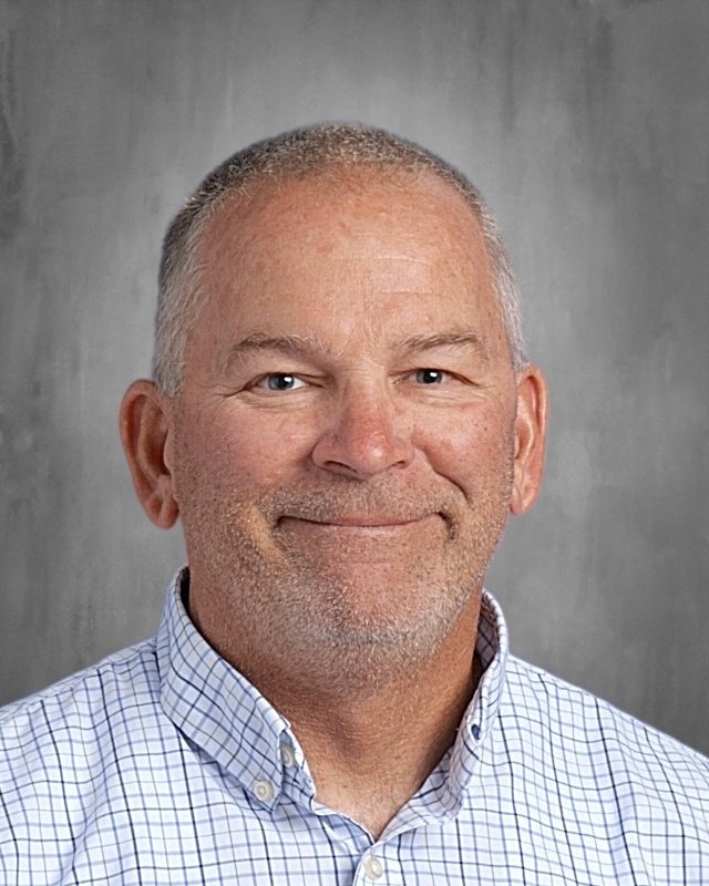 Portrait of a middle-aged man with short gray hair and a light beard, wearing a white checkered button-up shirt, smiling against a gray background.