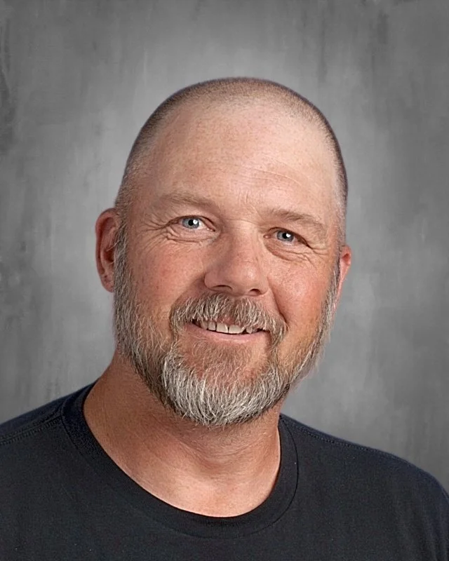 A smiling middle-aged man with a beard and short hair, wearing a black shirt, against a gray background.