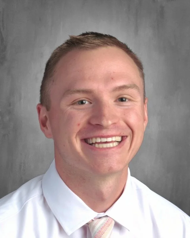 A smiling young man with short light brown hair in a white shirt and a light-colored tie, standing in front of a gray background.
