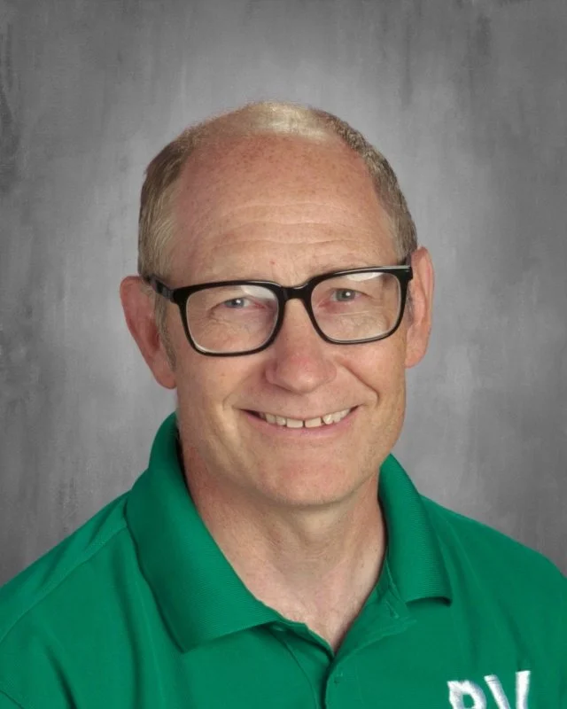 A middle-aged man with short, light-colored hair, wearing glasses and a green polo shirt, smiling in front of a gray background.