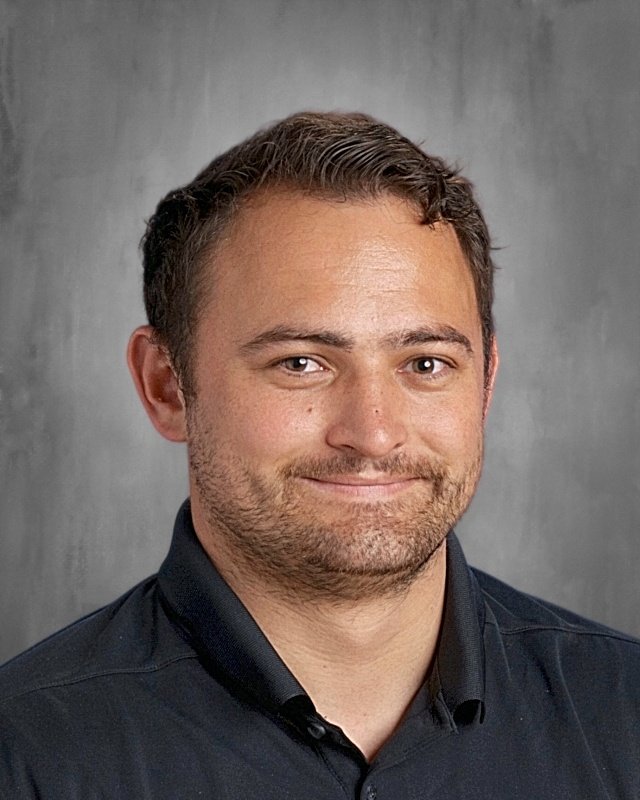 A young man with short brown hair and a beard, smiling, wearing a black collared shirt, against a grey background.
