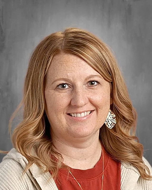 A smiling woman with wavy red hair wearing a beige jacket, red shirt, and large leopard print earrings against a gray background.