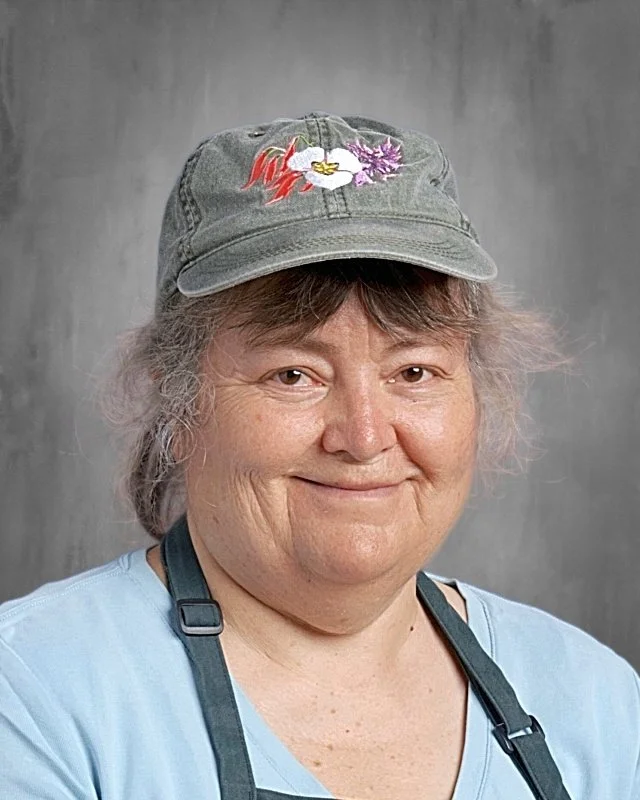 A smiling woman wearing a denim cap with an embroidered white flower and purple leaves, a light blue shirt, and a black apron against a gray background.