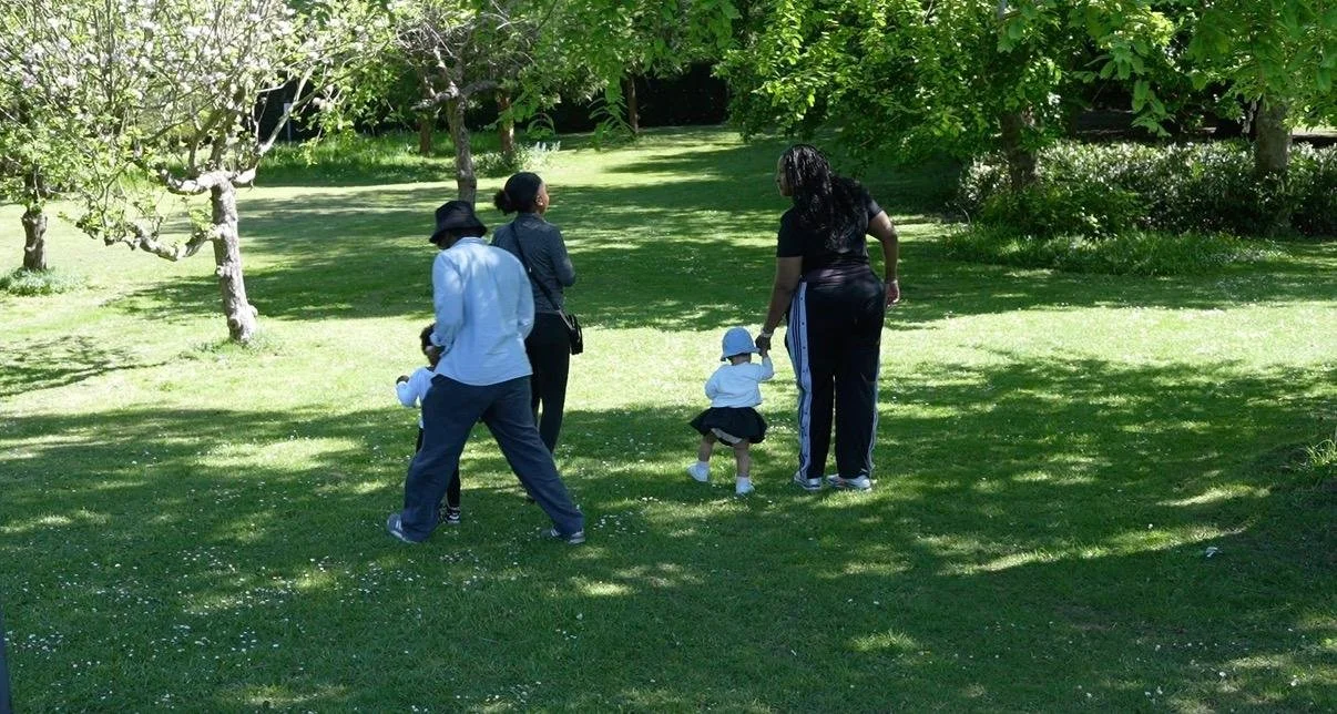 Four women and a young girl walking on a grassy park area under trees, some dappled shade, with trees and greenery in the background.
