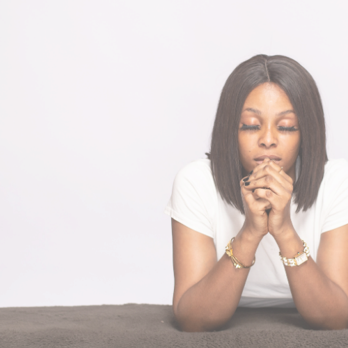 A woman with dark hair and makeup, wearing a white t-shirt, sitting with her hands clasped in front of her face, looking downward with eyes closed against a plain white background.