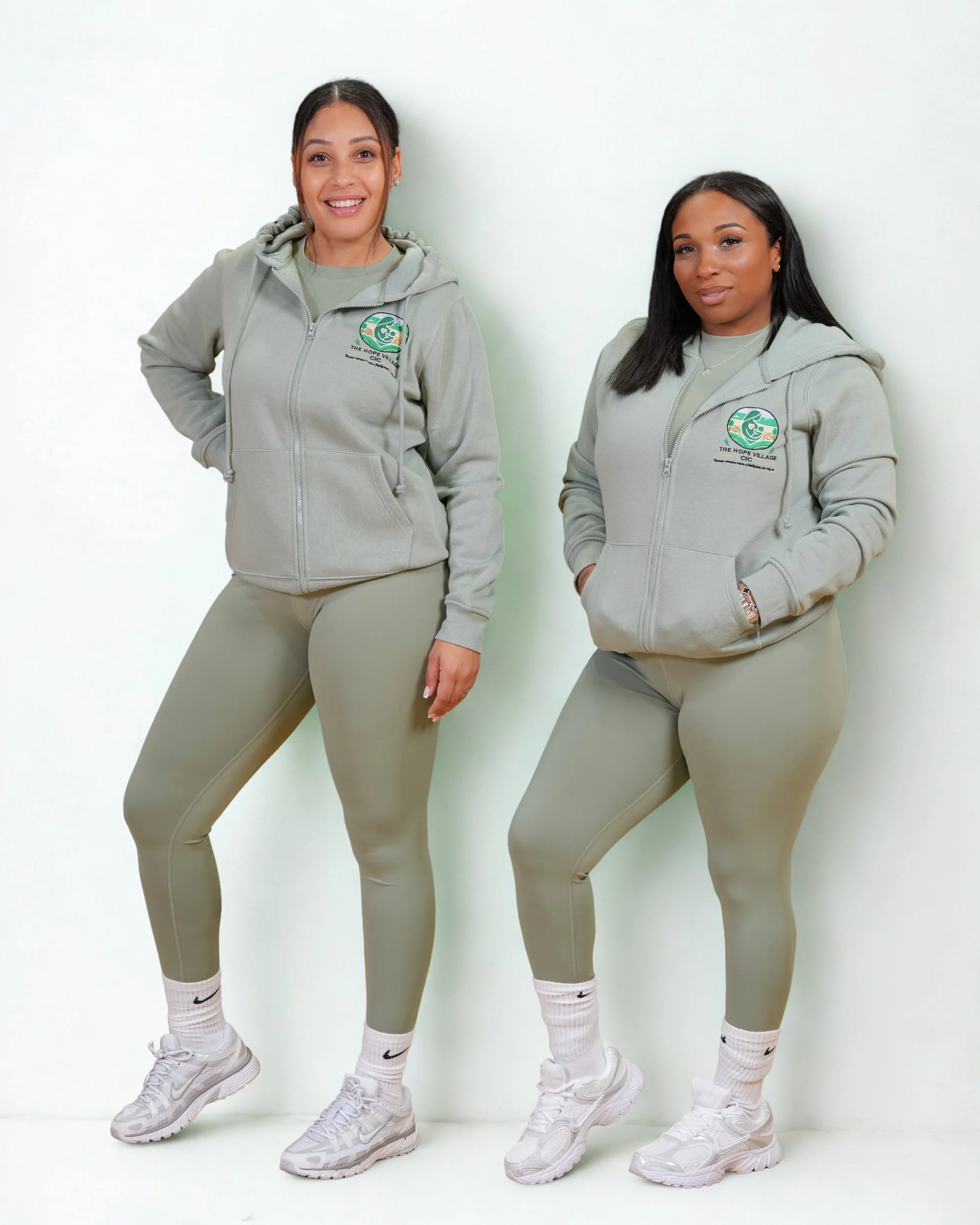 Two women standing against a white background, both wearing light gray hoodies with a logo and matching leggings and sneakers.