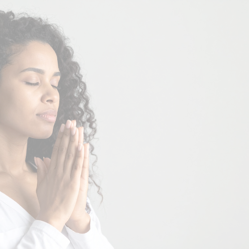 A woman with curly hair in a white top has her eyes closed and hands pressed together in prayer or meditation against a light background.