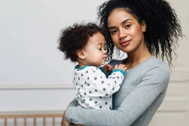 A woman holding a young child with curly hair in her arms, both with curly hair, standing against a plain light-colored background.