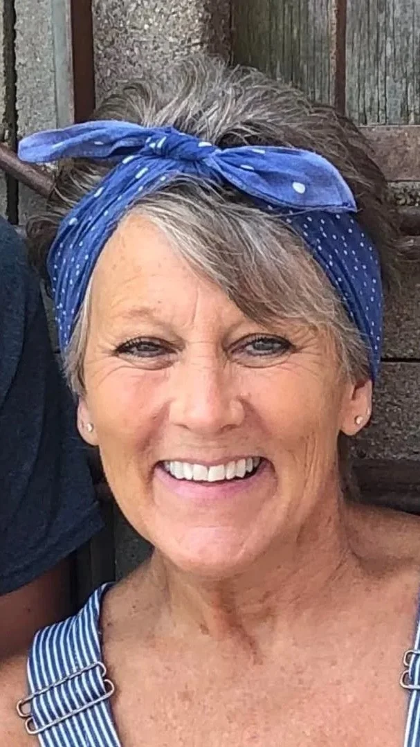 A smiling older woman with short gray hair wearing a blue polka-dotted bandana and striped sleeveless top.