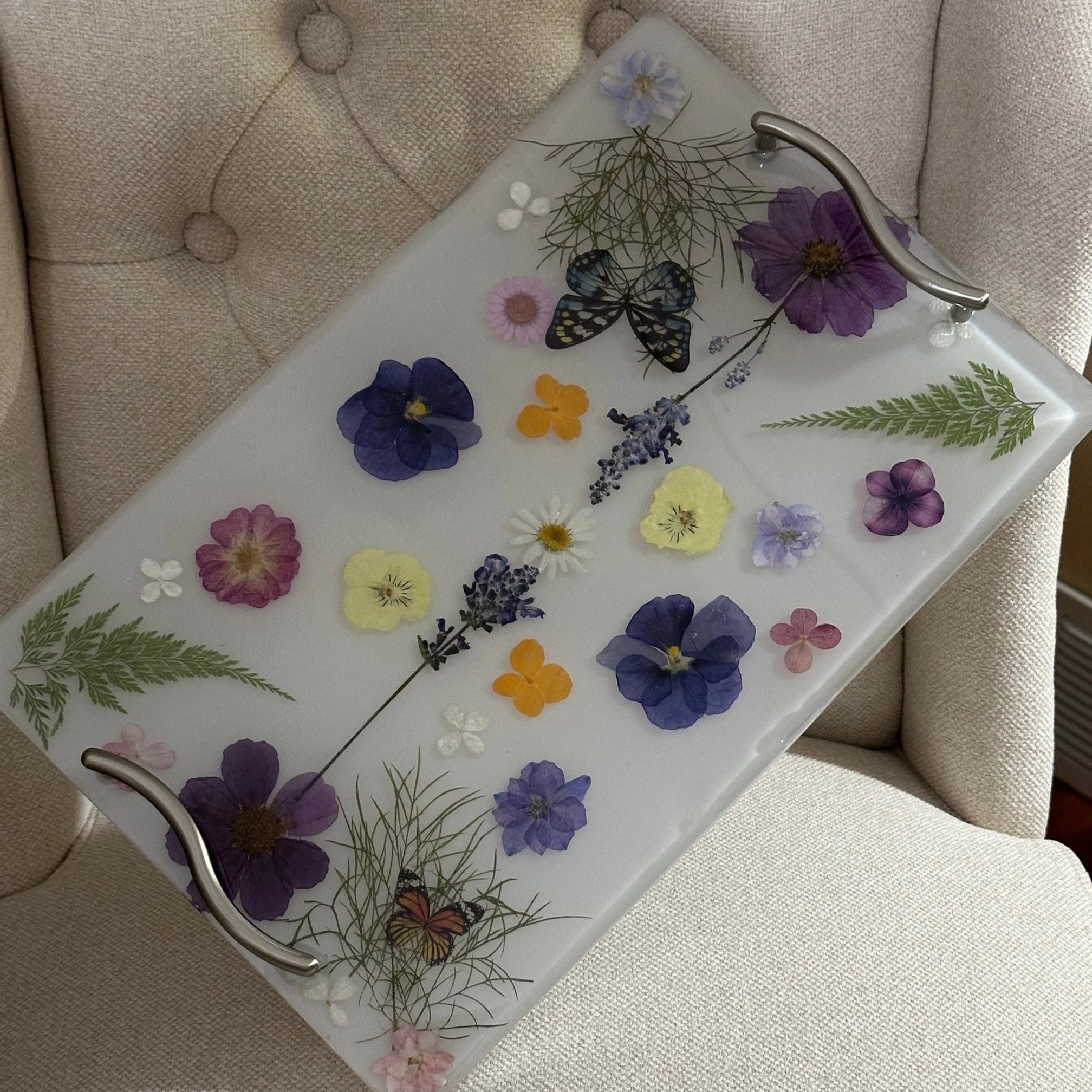 A glass tray with pressed flowers and butterflies, resting on a beige upholstered chair.