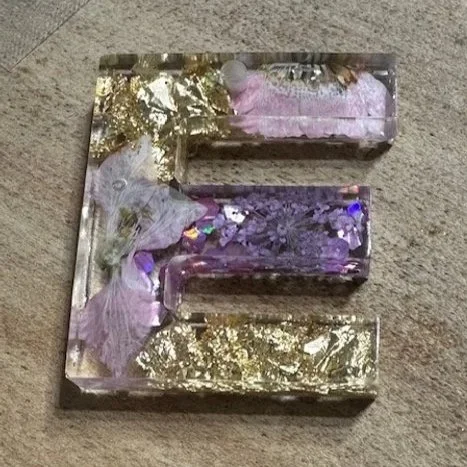 Decorative letter 'E' made of transparent resin with embedded purple flowers and gold leaf, placed on a beige carpet.
