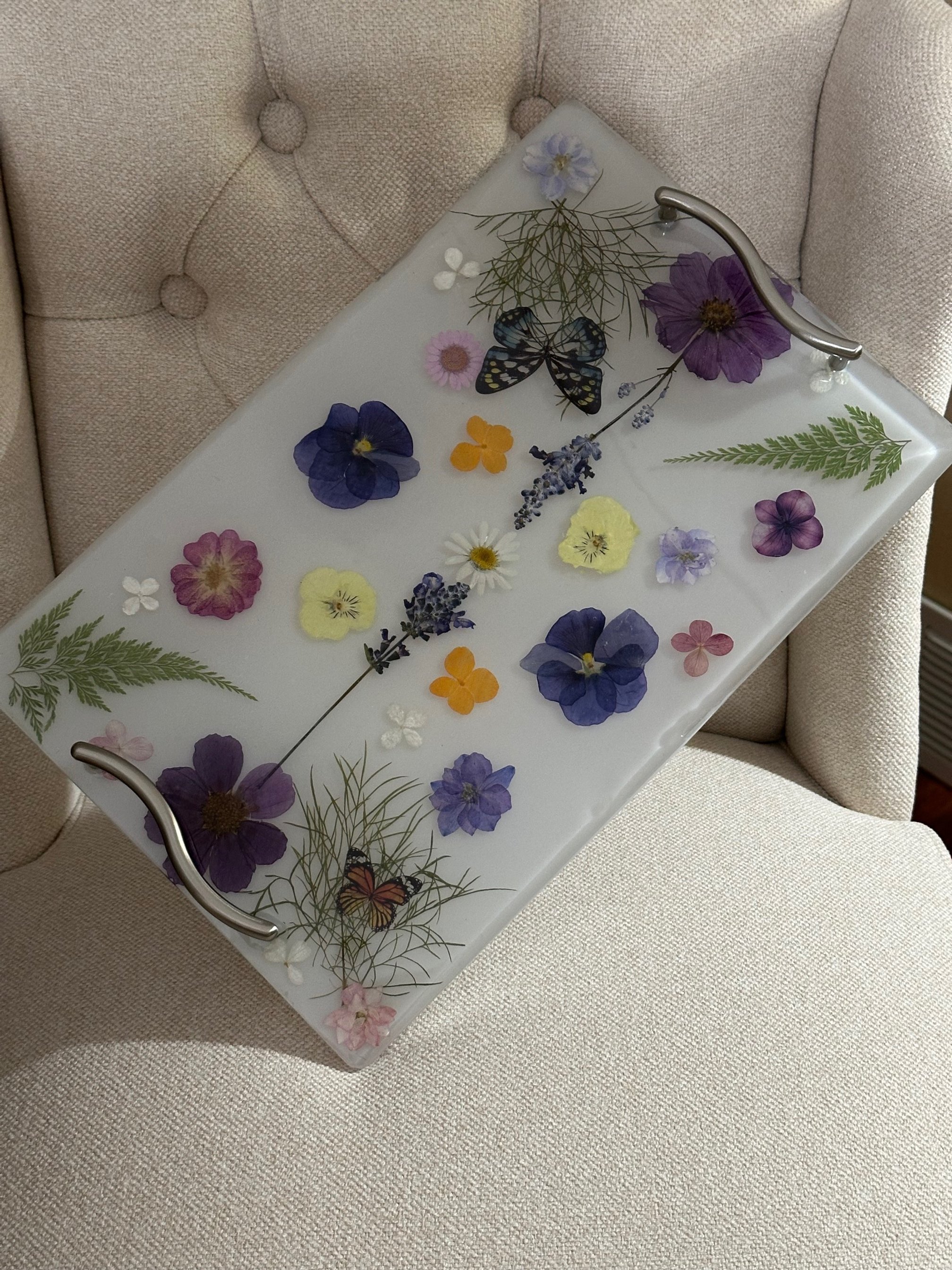 Decorative glass tray with pressed flowers and butterflies, on a beige fabric upholstered chair.