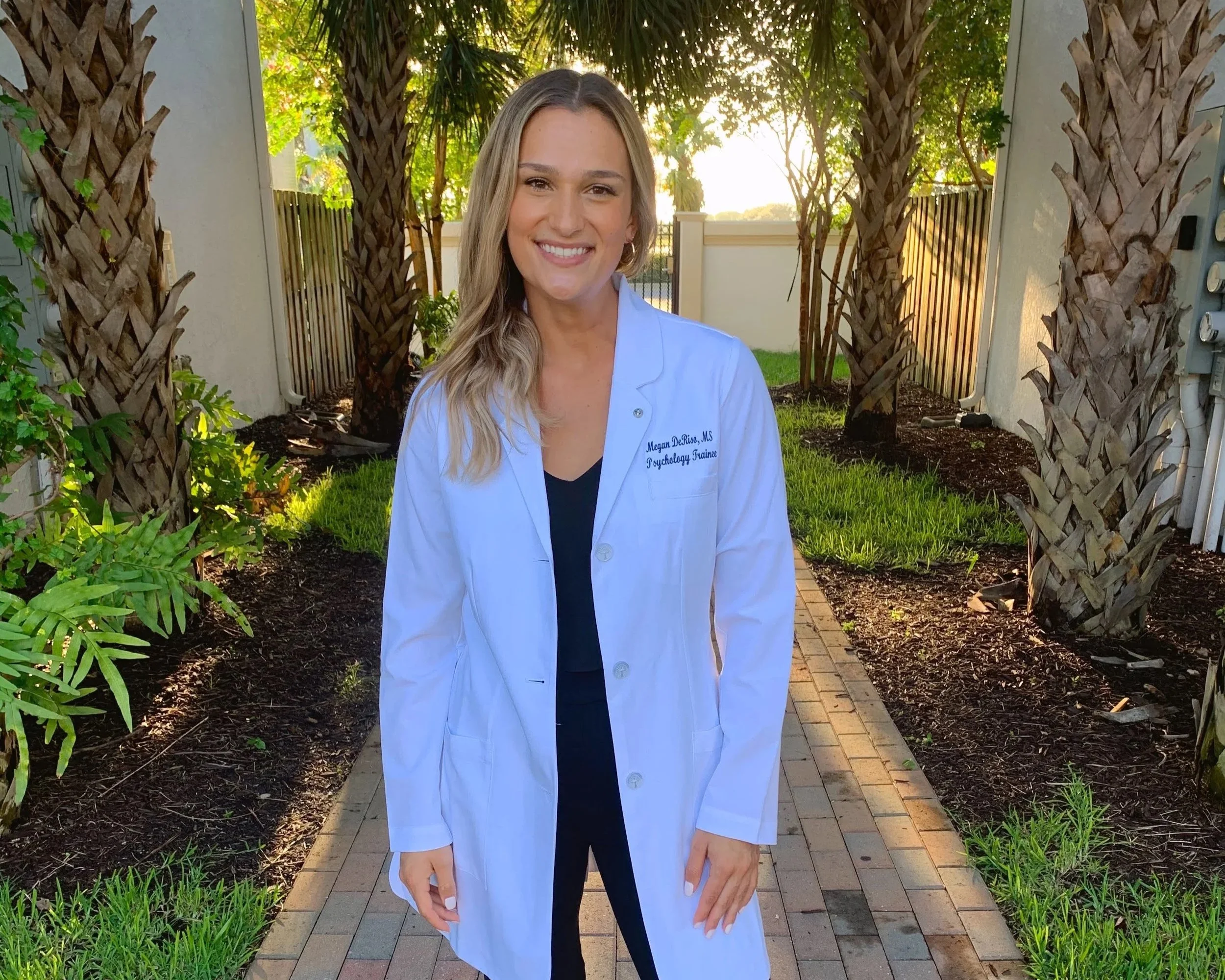 A woman in a white medical coat standing outdoors on a brick pathway, with palm trees and greenery in the background, smiling at the camera.