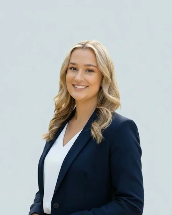 A smiling woman with blonde wavy hair wearing a navy blazer over a white blouse, standing against a light gray background.