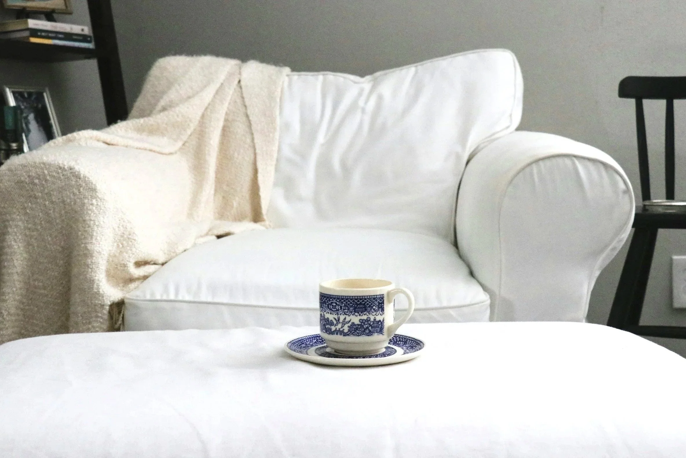 A white sofa with a cream blanket draped over one arm, a white cushion, and a black chair in the background. In the foreground, a white table holds a blue and white patterned teacup and saucer.