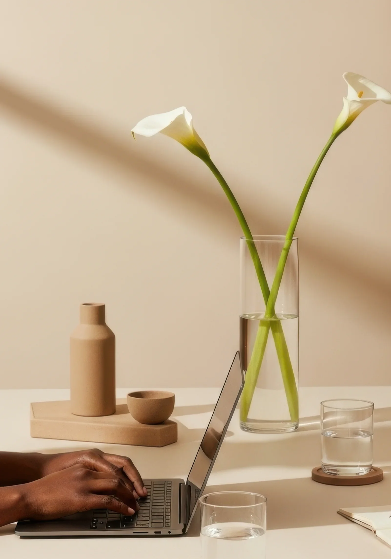 Person typing on a laptop at a white desk with a vase of water and two calla lilies, a beige vase, and decorative objects in neutral tones.