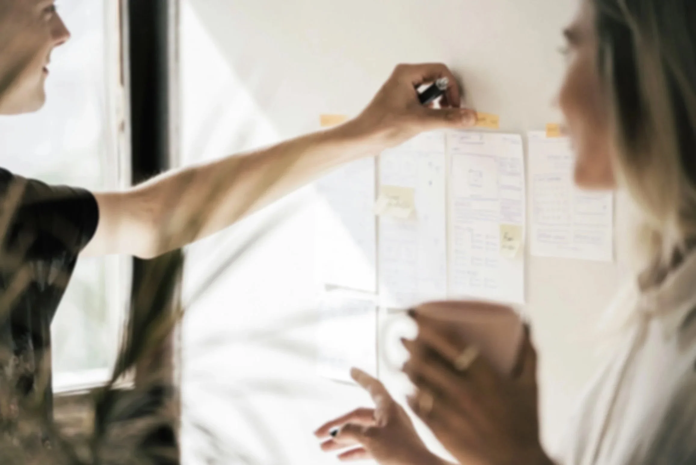 Two women discussing a planning board filled with notes and sketches, with one holding a pink mug and the other holding a black marker.