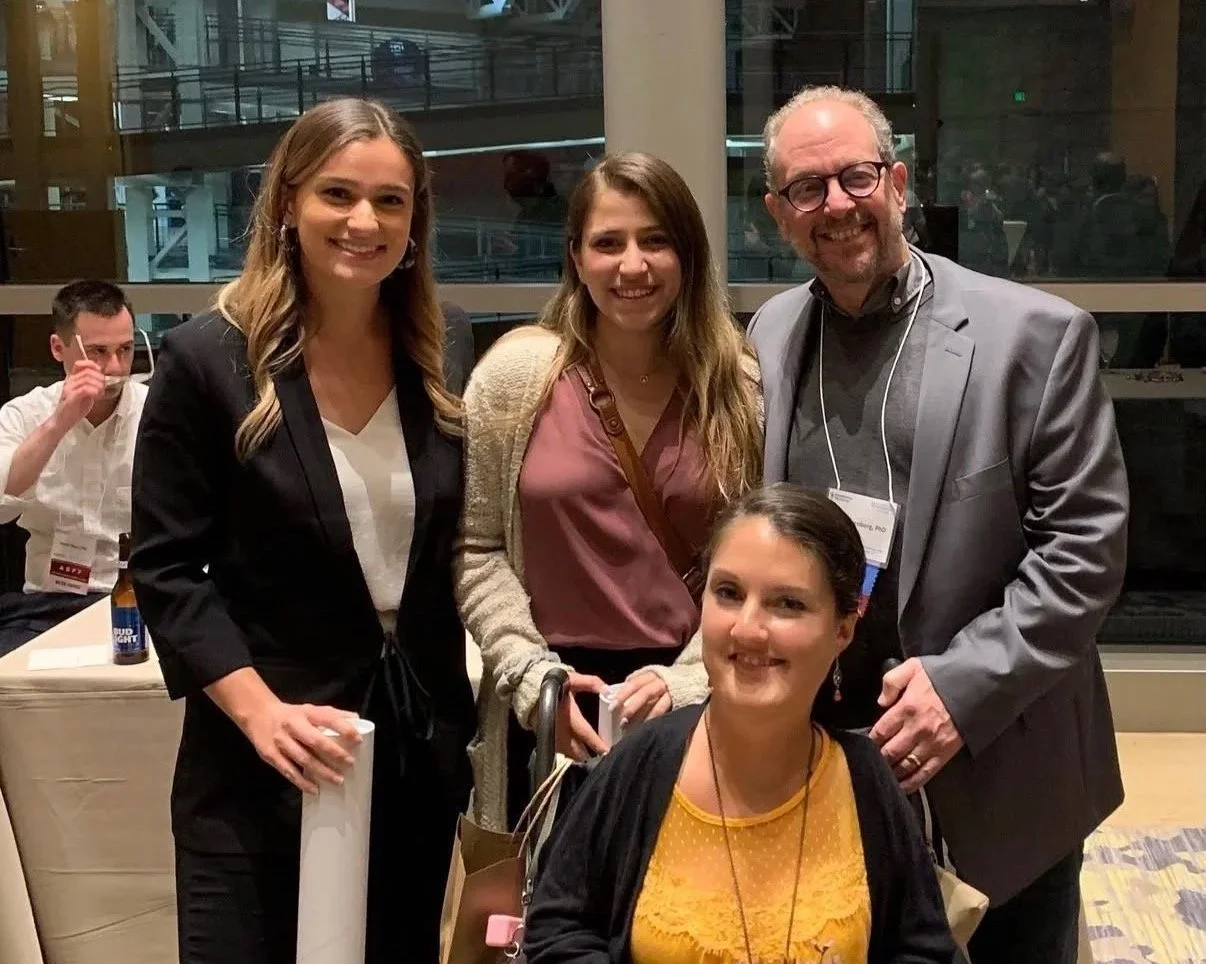 Group of five people smiling indoors at an event, with three women and two men. One woman is sitting, three are standing behind her, and one man is in the background wearing hearing aids. The background shows large windows and a modern interior.