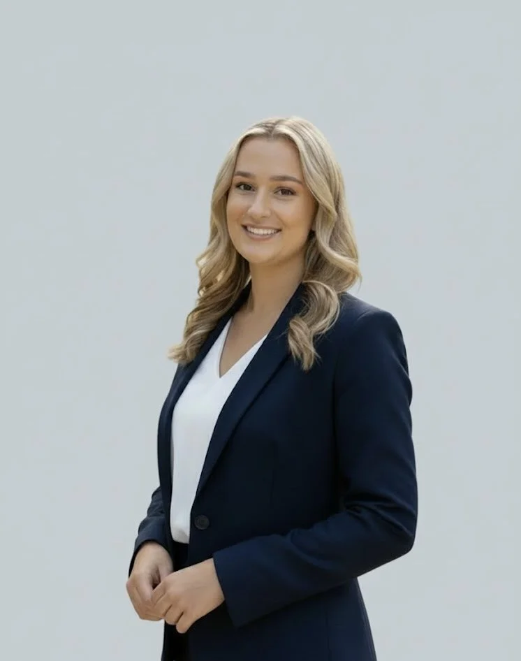 A young woman with blonde hair smiling, wearing a navy blazer and white blouse, standing against a plain light grey background.