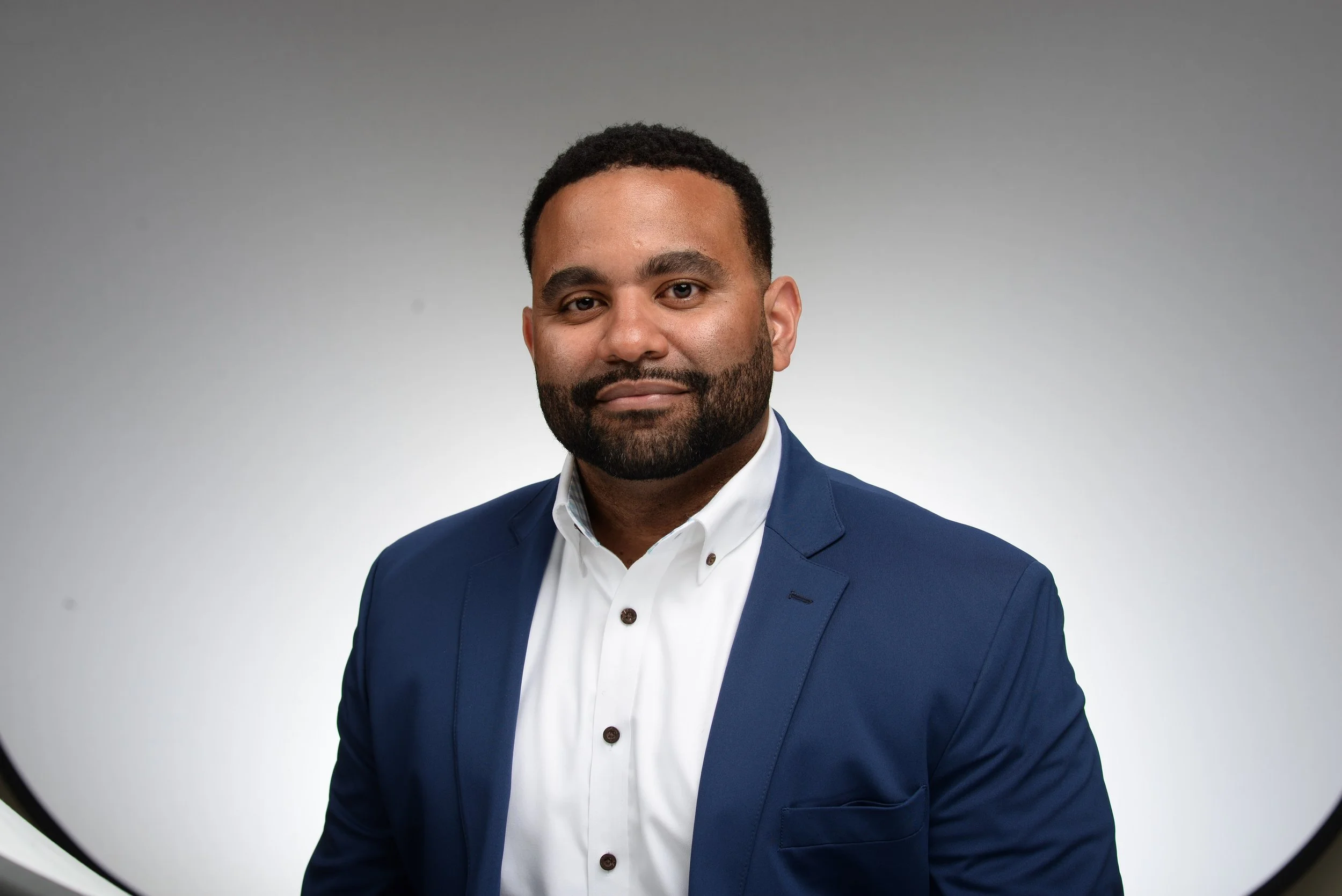 A man with dark hair and a beard in a blue suit and white shirt facing the camera with neutral expression, against a gray background.
