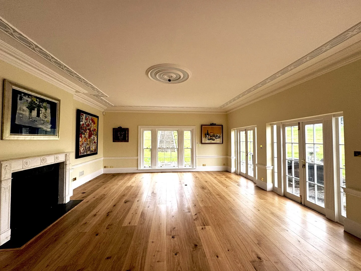 A spacious living room with light-colored walls, hardwood floors, a white fireplace mantle, framed artwork, large windows, and glass doors leading outside.