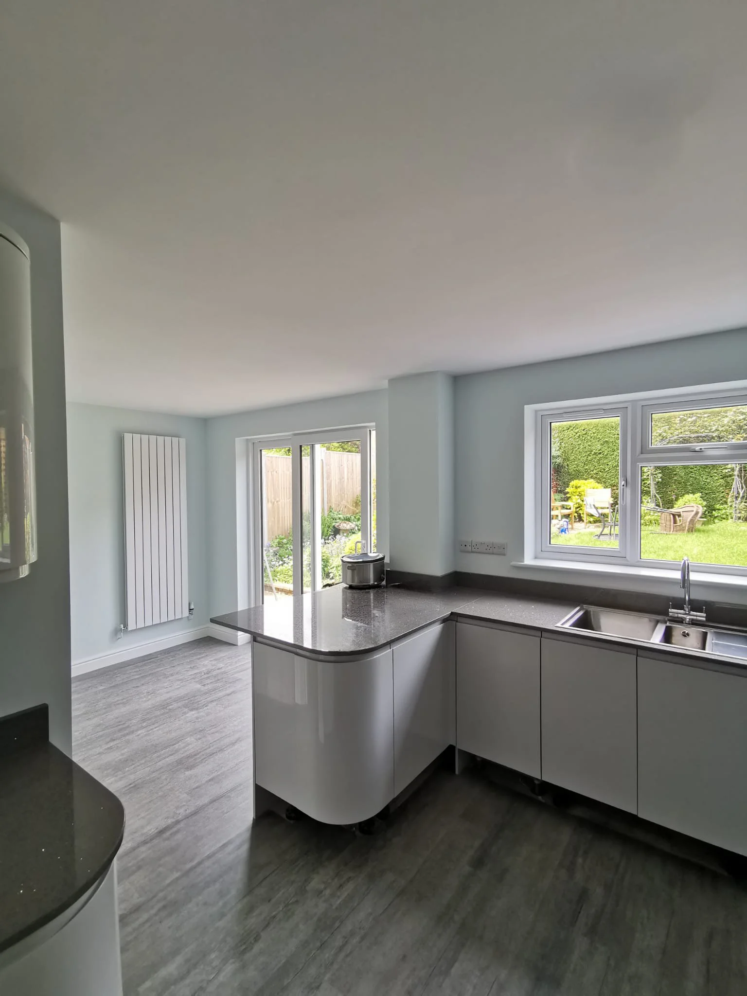 Modern kitchen with grey cabinets, stainless steel sink, large window overlooking a green garden, and sliding glass doors leading outside.