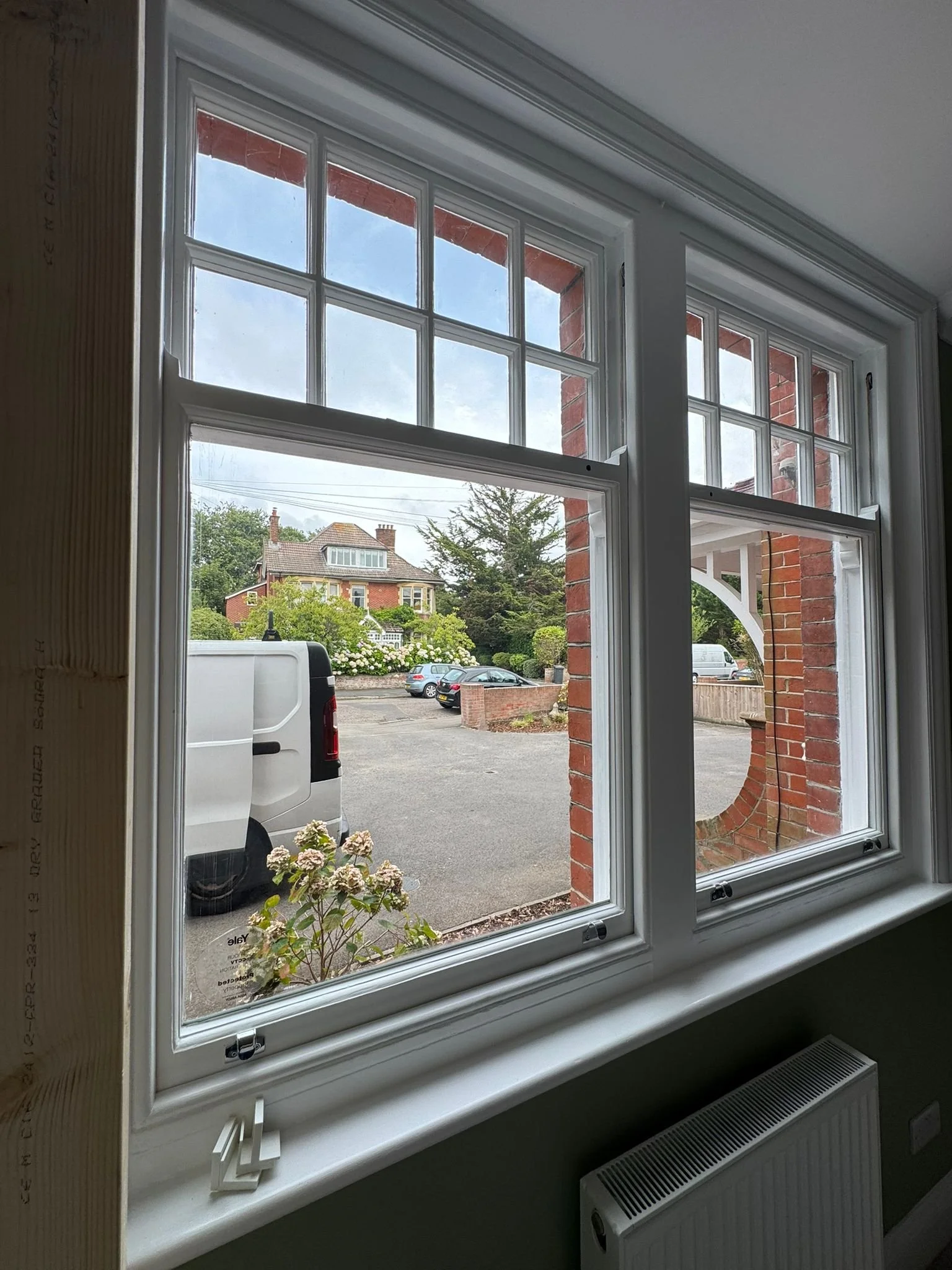 A view through a double casement window showing a parking lot, a white van, a flower bush, and a large house with a turret in the background.