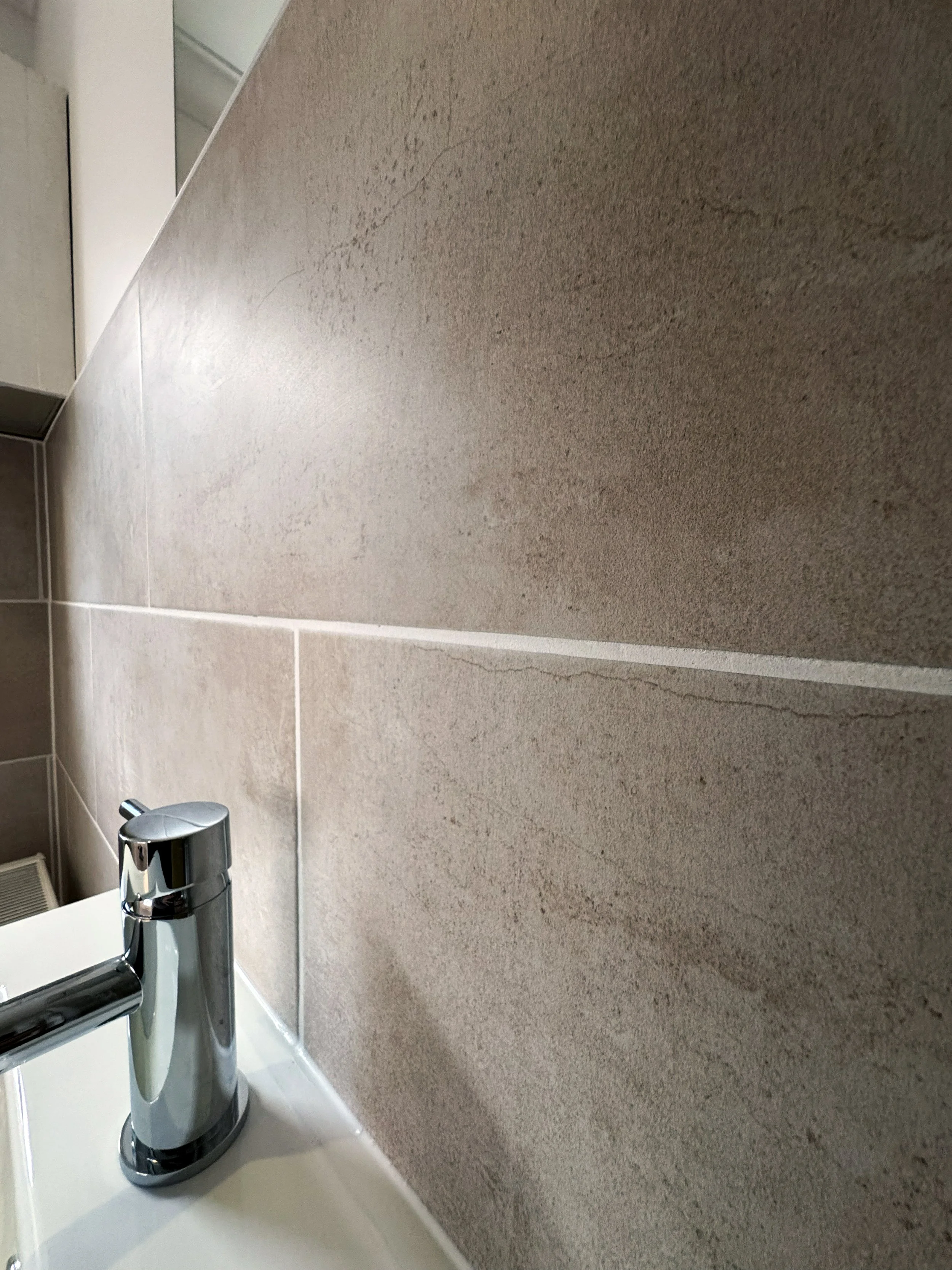 Close-up of a bathroom sink faucet with beige tiled wall in the background.