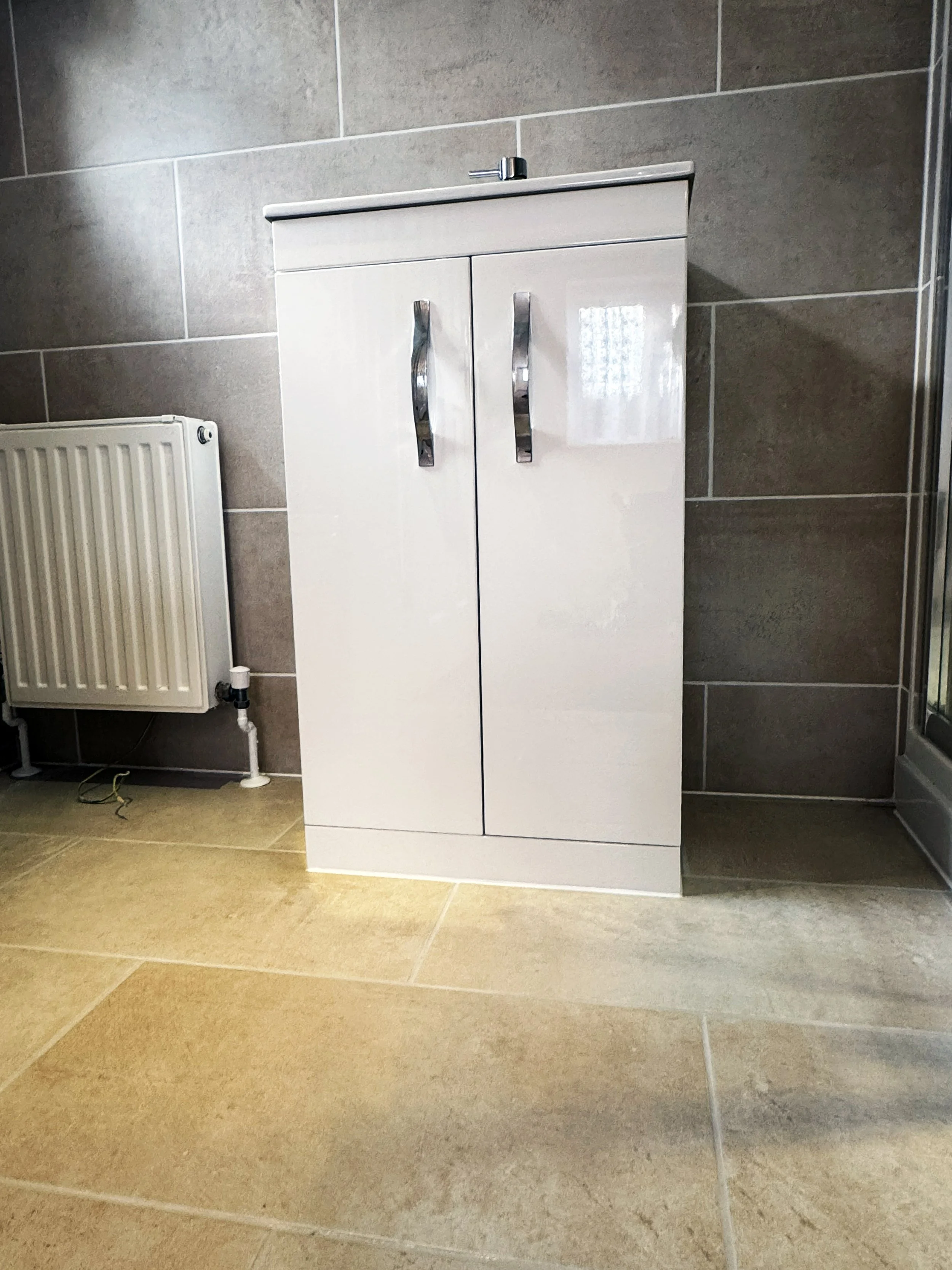 White bathroom cabinet with two doors and metallic handles in a tiled room with a radiator on the left and a glass shower door on the right.