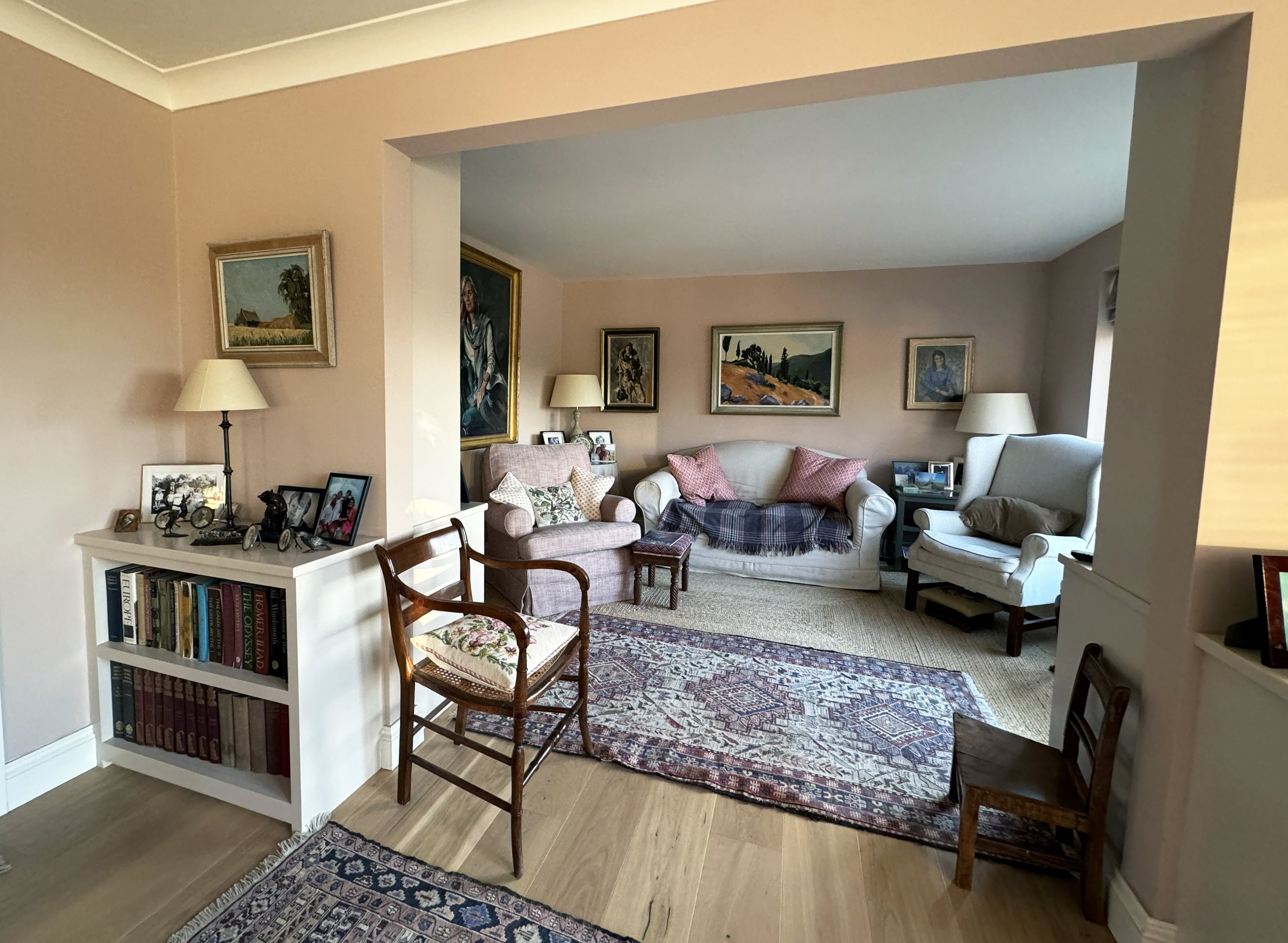 Living room with beige walls, a variety of framed paintings, a white couch with pink pillows and a plaid throw, a pink armchair, two high-back armchairs with lamps, a wood and fabric side table, a bookshelf with books and framed photos, and a multico