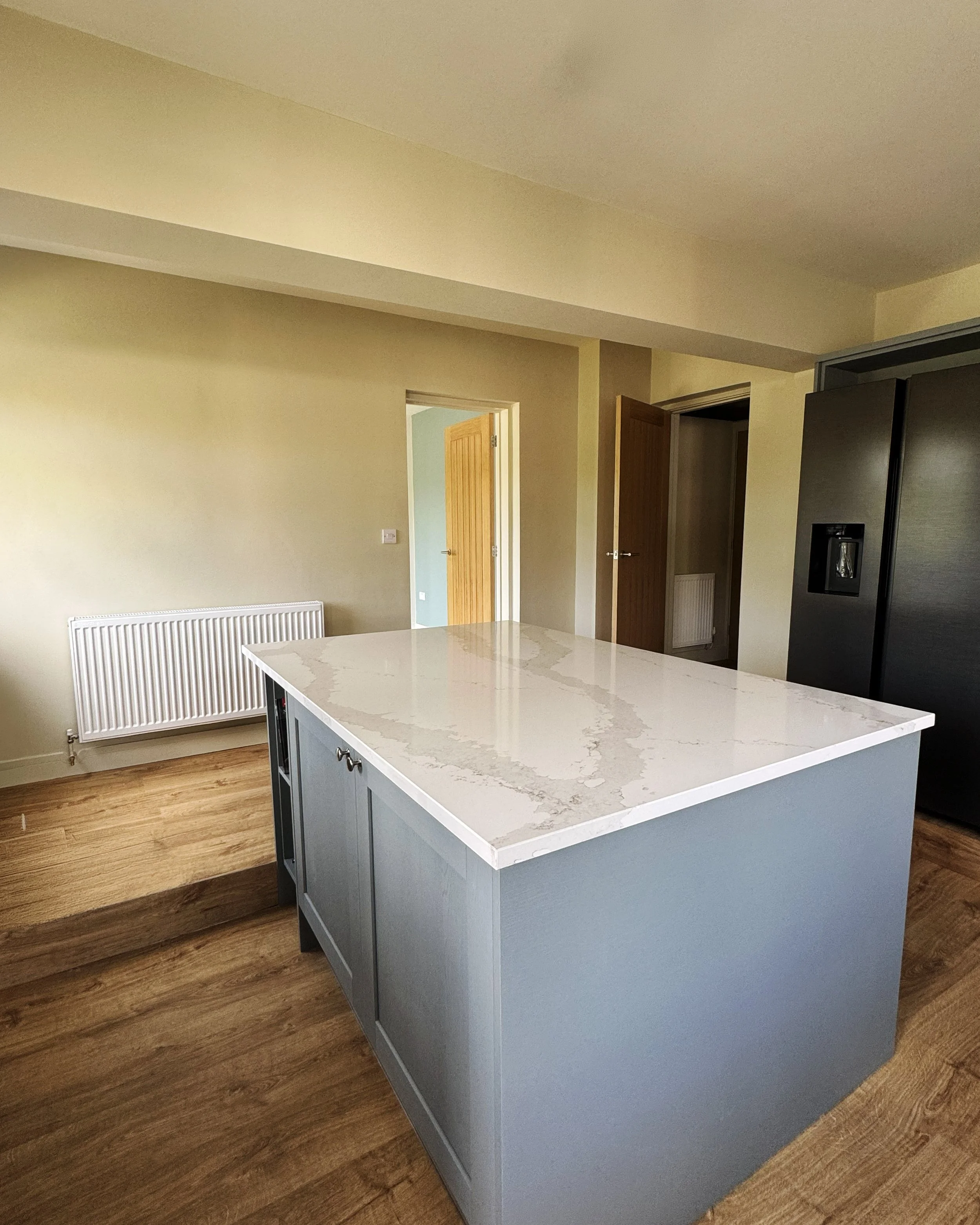 Modern kitchen with a marble countertop island, light green walls, and wooden flooring. There is a black refrigerator and open doors leading to other rooms.