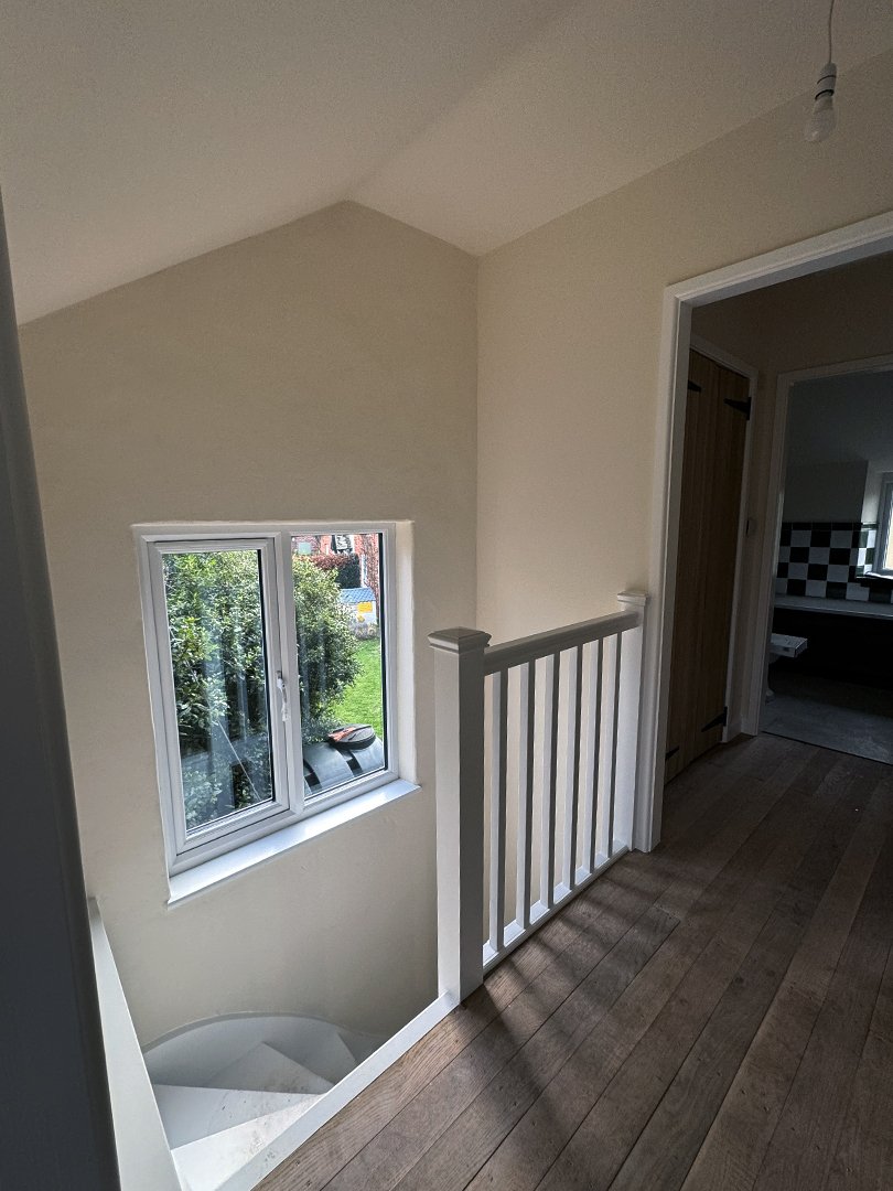 Interior view of a stairs landing with a window overlooking greenery, a white railing, and an open doorway leading to a kitchen with black-and-white checkered backsplash.