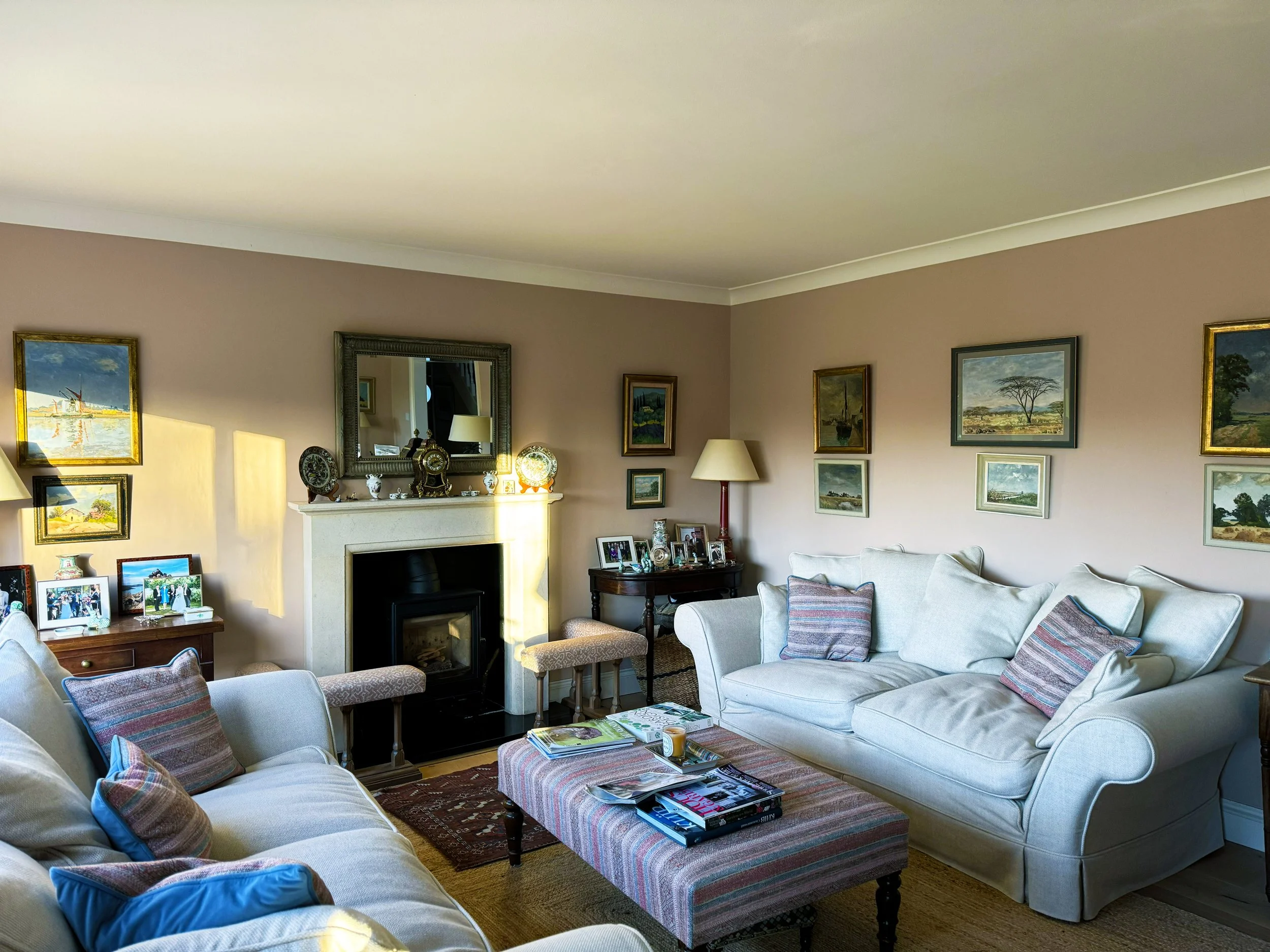 Living room with beige walls, three sofas with striped cushions, a fireplace with a mirror above, several framed landscape paintings, and a coffee table with magazines and candles.